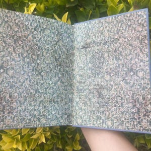 May include: Open book with a blue cover and patterned pages. The pages have a teal background with a white floral design. The book appears to be open, revealing the inside pages with handwritten text.