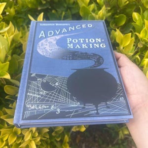 May include: A blue book titled "Advanced Potion-Making" with a black cauldron illustration. The book's cover features the author's name, a swirling design, and various symbols. The book is held against a backdrop of green foliage.