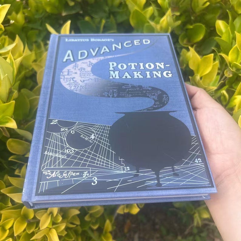 May include: A blue book titled "Advanced Potion-Making" with a black cauldron illustration. The book's cover features the author's name, a swirling design, and various symbols. The book is held against a backdrop of green foliage.