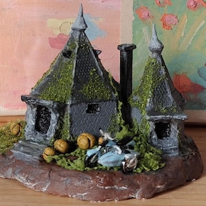 May include: A miniature gray stone cottage with two pointed roofs, a tall chimney, and green moss accents. The structure sits on a brown base with small round tan objects and a light blue motorcycle with a sidecar parked in the front.