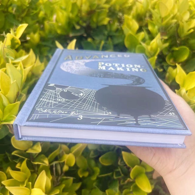 May include: A blue hardcover book titled "Advanced Potion Making" with a cauldron illustration. The cover features a moon and scientific diagrams. The book is held against a backdrop of green foliage.