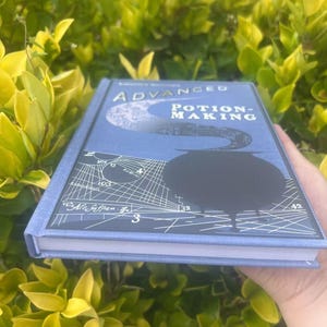 May include: A hardcover book titled "Advanced Potion-Making" with a blue cover. The cover features a black cauldron, a crescent moon, and white geometric designs. The book is held against a backdrop of green foliage.