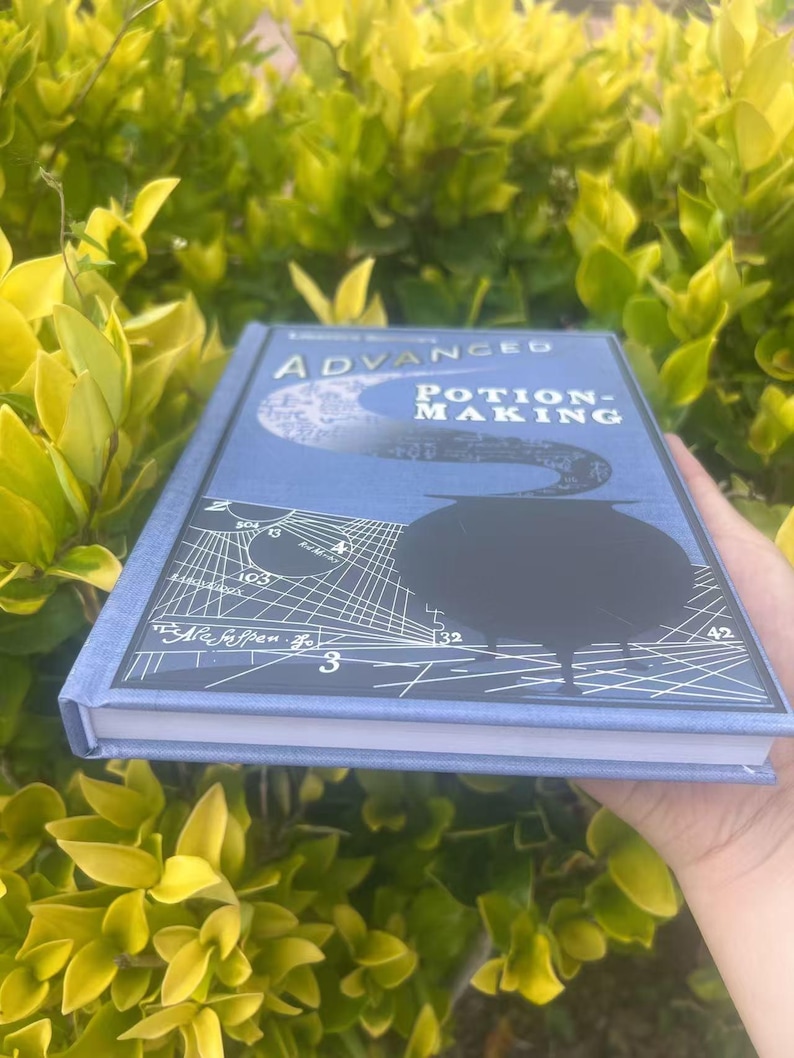 May include: A hardcover book titled "Advanced Potion-Making" with a blue cover. The cover features a black cauldron, a crescent moon, and white geometric designs. The book is held against a backdrop of green foliage.