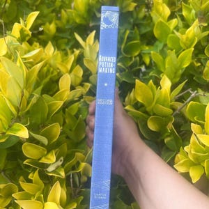 May include: A blue book titled "Advanced Potion-Making" with white text and geometric designs. The book's spine is held against a backdrop of green and yellow foliage. The book is a second edition by Libatius Borage.