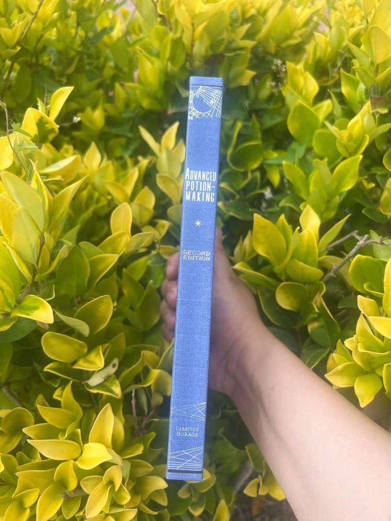 May include: A blue book titled "Advanced Potion-Making" with white text and geometric designs. The book's spine is held against a backdrop of green and yellow foliage. The book is a second edition by Libatius Borage.