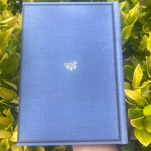 May include: A blue book with a textured cover and a decorative emblem in the lower center. The book has a dark border and is set against a backdrop of green foliage.