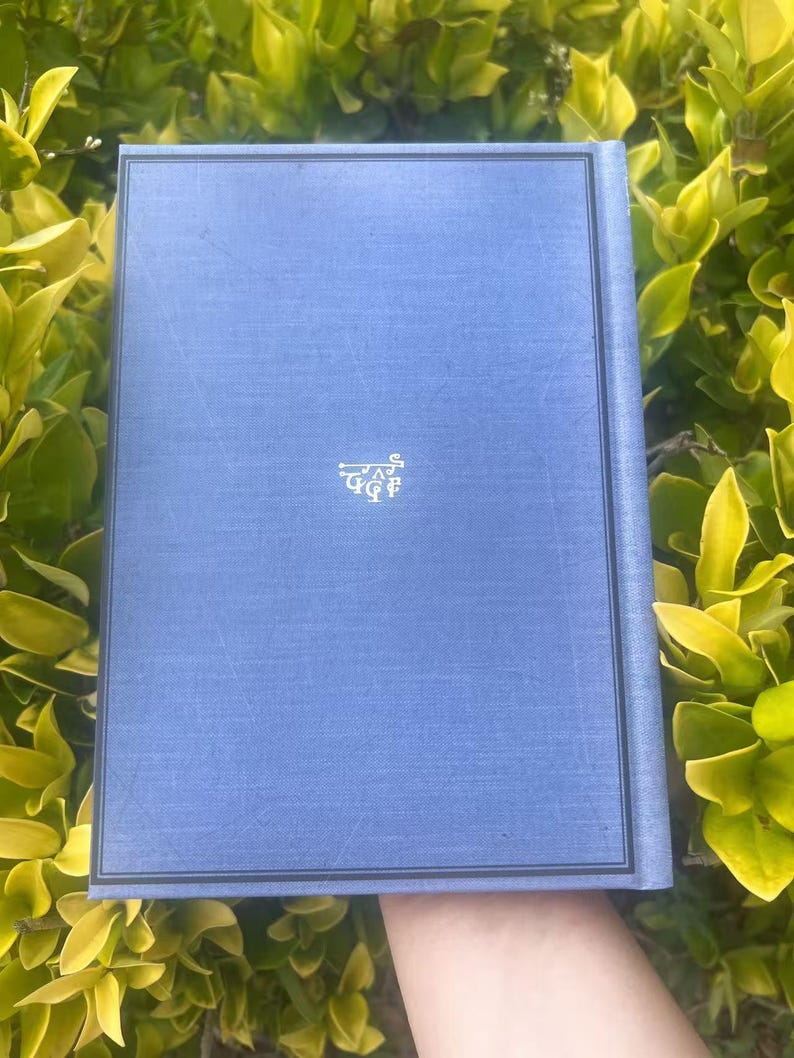 May include: A blue book with a textured cover and a decorative emblem in the lower center. The book has a dark border and is set against a backdrop of green foliage.