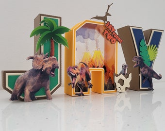May include: A colorful cardboard letter set spelling "JAY TURNS TWO" with dinosaur and volcano illustrations. The letters are decorated with green palm trees, brown rocks, and green leaves.