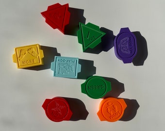 May include: Seven colorful, 3D printed game pieces with various words and symbols. The pieces are shaped like triangles, squares, and circles. The words on the pieces include "More", "Play-Doh", "Green", "Want", "No", "Yes", and "All Done".
