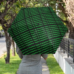 Puede incluir: Un paraguas negro con un patrón verde repetido del texto "Negative Type O Negative Type O" en el exterior. El paraguas está siendo sostenido por una persona que lleva un vestido de cuadros grises y negros.