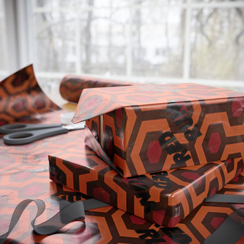 May include: Two wrapped gift boxes with a repeating geometric pattern in orange, brown, and red. The boxes are stacked on top of each other. The words "RED" are printed on the side of the top box. A pair of scissors and wrapping paper are also visible.