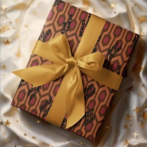 May include: A wrapped gift box with a gold satin ribbon and bow. The wrapping paper features a geometric pattern in brown and burgundy, with the words "RED RUM" printed on it. The gift is presented on a white satin surface with gold star-shaped confetti.