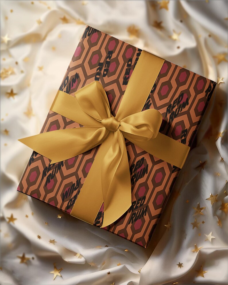 May include: A wrapped gift box with a gold satin ribbon and bow. The wrapping paper features a geometric pattern in brown and burgundy, with the words "RED RUM" printed on it. The gift is presented on a white satin surface with gold star-shaped confetti.
