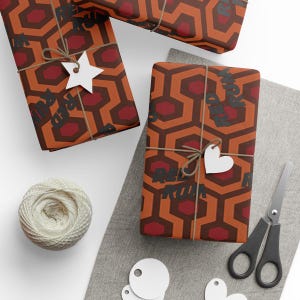 May include: Gift-wrapped presents with a repeating geometric pattern in shades of orange and red. The wrapping paper features the text "RED RUM" in black. Each gift is tied with twine and a white paper tag. A ball of twine, scissors, and blank tags are also visible.
