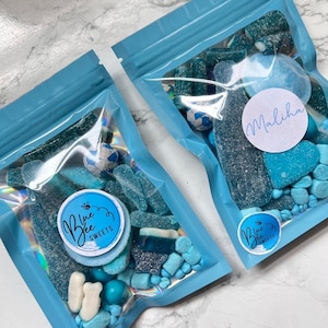 May include: Two blue cellophane bags filled with blue and white sweets. The bags have a blue zip closure. One bag has a round sticker that says "Blue Bee Sweets". The other bag has a sticker that says "Maliha".