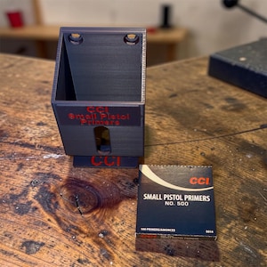 May include: A dark gray 3D-printed plastic dispenser for CCI Small Pistol Primers with red lettering on the front and a matching box of CCI Small Pistol Primers No. 500 positioned next to it. The dispenser features two mounting holes at the top.
