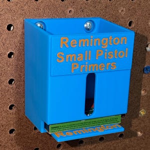 May include: A bright blue plastic wall-mounted dispenser featuring the embossed text Remington Small Pistol Primers on the front. The unit includes a vertical cutout window and a small bottom shelf holding a box of Remington ammunition.