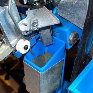 May include: Close-up of a blue reloading press with a metal shell holder and a white plastic pulley. The press has a metal arm and a blue plastic tray.
