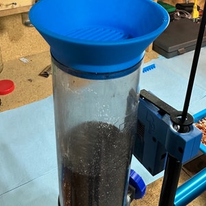 May include: A blue plastic funnel sits atop a clear glass cylinder filled with black powder. The cylinder is attached to a blue and black reloading press.