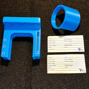 May include: Bright blue 3D-printed firearm accessories, including a frame and a circular component. Two white cards with fillable fields for firearm specifications are also present. The cards include fields for caliber, bullet type, and other details.