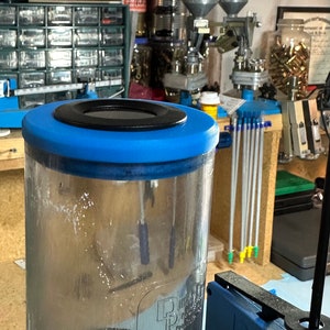 May include: A clear, cylindrical container with a blue lid and a black center, filled with dark granules. The container is part of a reloading setup, with other tools and components visible in the background.