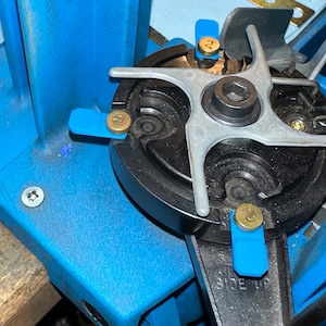 May include: Close-up of a blue reloading press with a black metal star-shaped die holder. The die holder has four numbered positions, each with a blue and gold screw. The text "SIDE UP" is visible on the press.