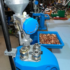 May include: A blue reloading press with a silver hopper and a blue dial. The press is on a blue surface with a tray of copper-colored bullets.