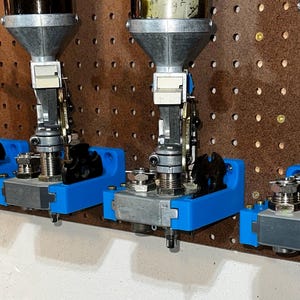 May include: Four reloading presses mounted on a brown pegboard. Each press has a silver funnel, a blue base, and various metal components. The presses are designed for reloading ammunition.