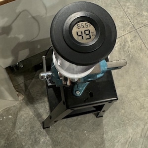 May include: A black device with a digital display showing 65.5°F and 49%. The device is mounted on a metal and black stand, secured by a blue vise. The setup is on a concrete floor.