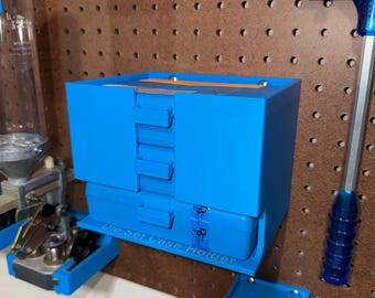 May include: A bright blue Die Set Case Holder is mounted on a brown pegboard. The holder has a rectangular shape with multiple compartments and the text "Die Set Case Holder" is visible. Other reloading tools are also visible.