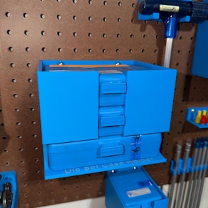 May include: Bright blue die set case holder mounted on a brown pegboard. The holder has a box shape with multiple compartments and the text "Die Set Case Holder". Other blue tools are also mounted on the pegboard.