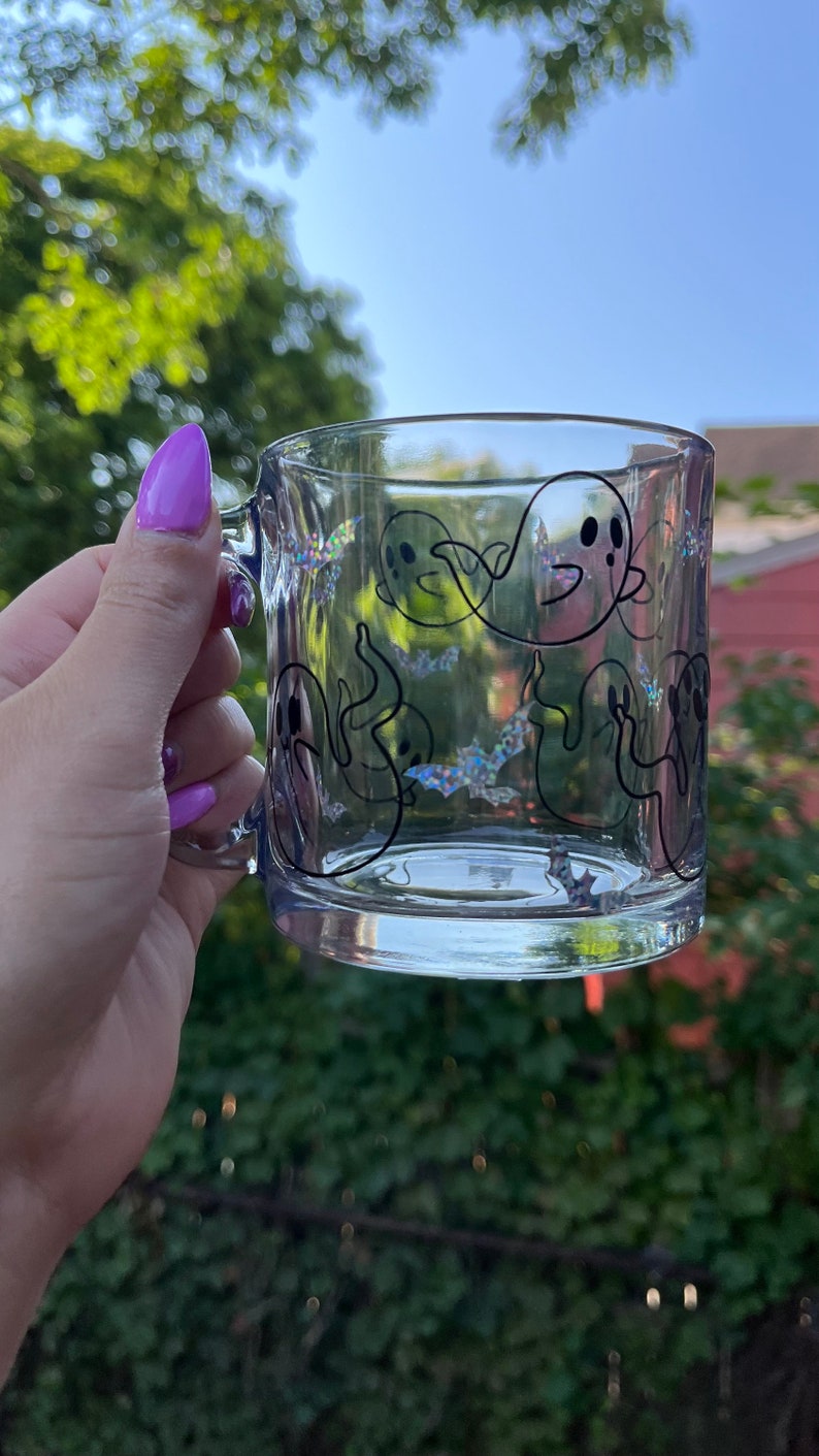 May include: A clear glass mug with a black outline of ghosts and bats. The mug has a glittery iridescent finish.