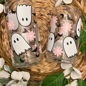 May include: Two glass tumblers with a whimsical Halloween design. The tumblers feature pink flowers, white ghosts, black bats, and crescent moons.