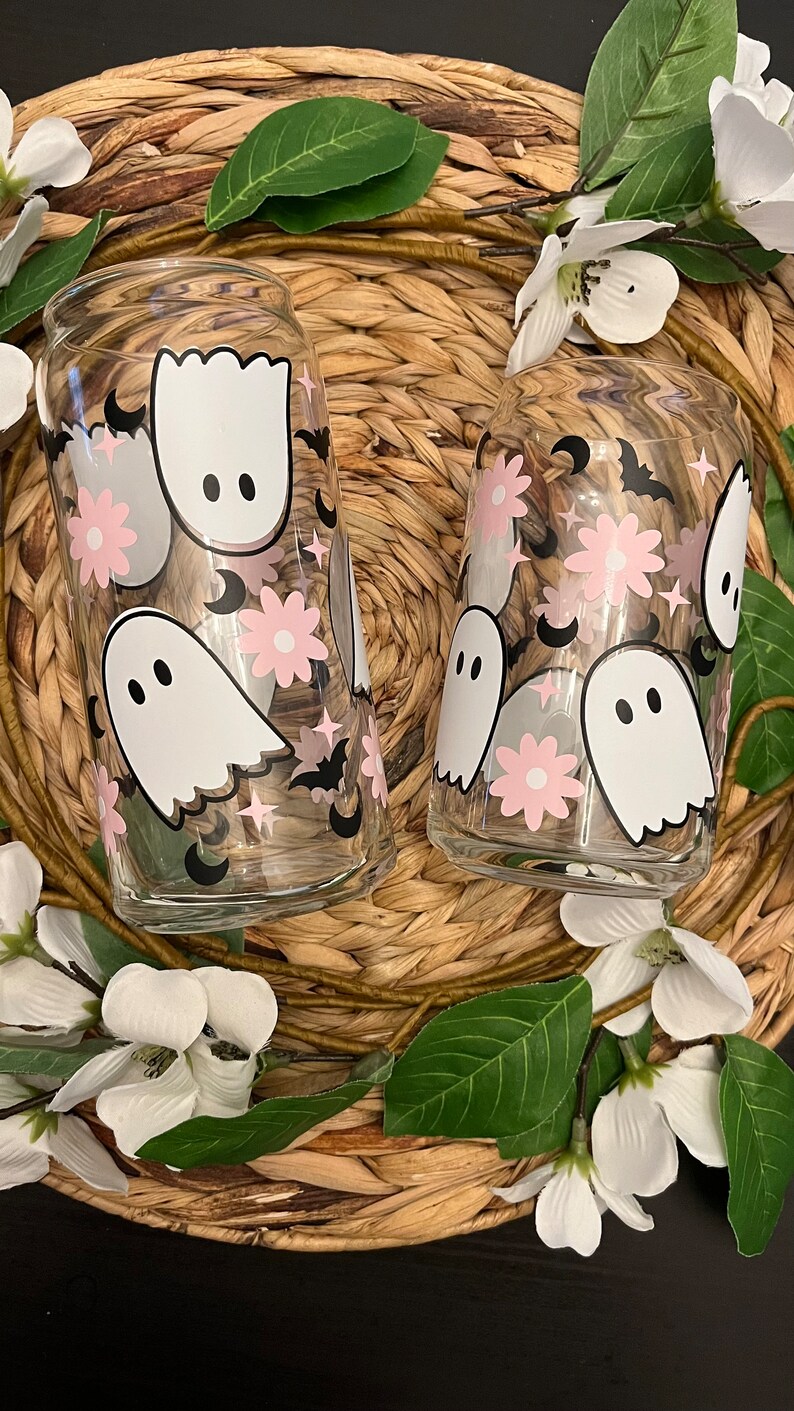 May include: Two glass tumblers with a whimsical Halloween design. The tumblers feature pink flowers, white ghosts, black bats, and crescent moons.