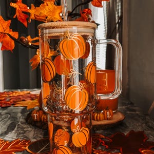 Könnte beinhalten: Ein klares Glas mit Henkel und Bambusdeckel, verziert mit orangefarbenen Kürbismotiven. Das Glas ist mit einem Getränk und einem Strohhalm gefüllt. Herbstblätter und dekorative Kürbisse umgeben das Glas.