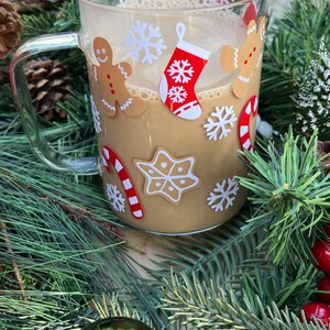 May include: A clear glass mug with a handle, filled with a brown beverage. The mug is decorated with Christmas-themed stickers, including gingerbread men, snowflakes, candy canes, and a stocking.