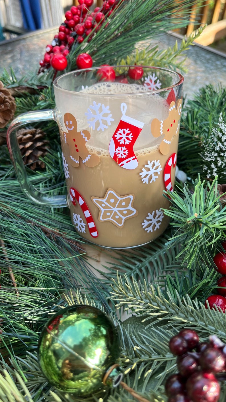 May include: A clear glass mug with a handle, filled with a brown beverage. The mug is decorated with Christmas-themed stickers, including gingerbread men, snowflakes, candy canes, and a stocking.