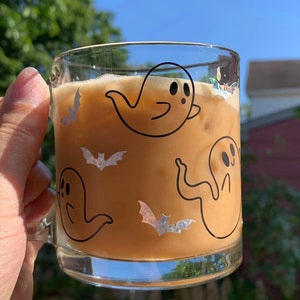 May include: A glass mug with a brown beverage inside. The mug has a white background with black ghost and bat illustrations.