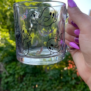 May include: A clear glass mug with a black and white ghost design. The mug has a handle and is filled with a clear liquid.
