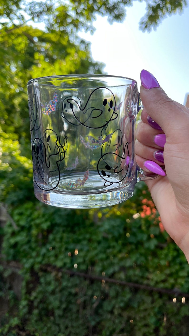May include: A clear glass mug with a black and white ghost design. The mug has a handle and is filled with a clear liquid.