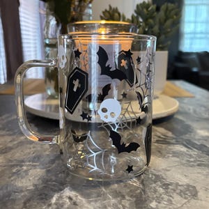 Pode incluir: Uma caneca de vidro transparente com um design de Halloween em preto e branco. O design apresenta morcegos, caveiras, teias de aranha e estrelas.