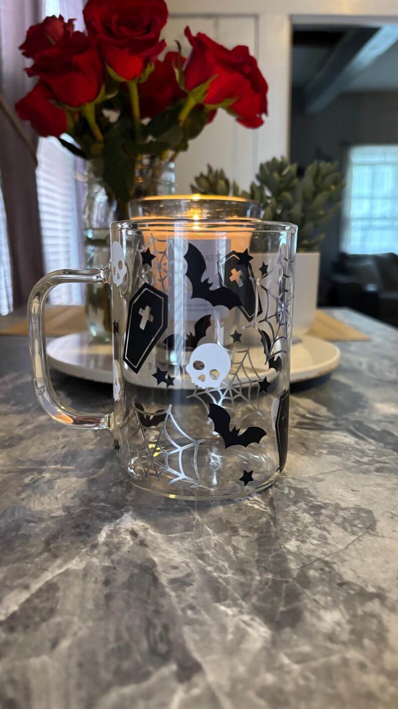 Pode incluir: Uma caneca de vidro transparente com um design de Halloween em preto e branco. O design apresenta morcegos, caveiras, teias de aranha e estrelas.