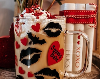 May include: A clear glass mug filled with a creamy beverage, topped with whipped cream and red sprinkles. The mug is decorated with black lips, red hearts, and a bat. The words "Spooky Babe", "HUGS", and "XOXO" are visible.