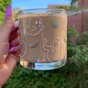 May include: A clear glass mug with a brown beverage inside. The mug has a white outline of ghosts and bats with iridescent glitter accents.