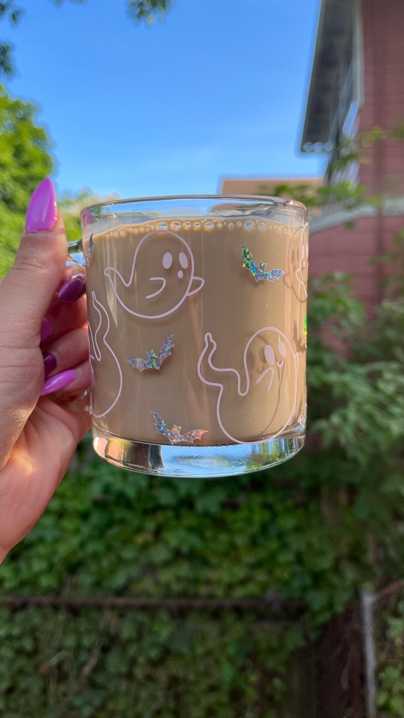 May include: A clear glass mug with a brown beverage inside. The mug has a white outline of ghosts and bats with iridescent glitter accents.