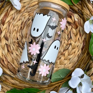 May include: A clear glass reusable water bottle with a bamboo lid and a black straw. The bottle has a whimsical design featuring white ghosts, pink flowers, black bats, and silver stars.