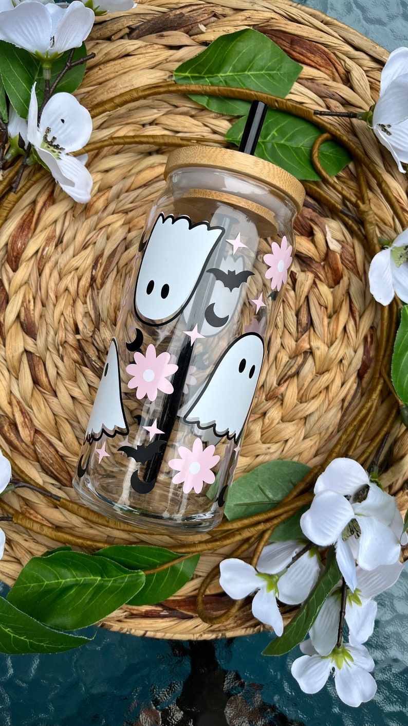 May include: A clear glass reusable water bottle with a bamboo lid and a black straw. The bottle has a whimsical design featuring white ghosts, pink flowers, black bats, and silver stars.