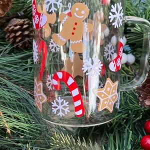 May include: A clear glass mug with a festive Christmas design. The mug features a gingerbread man, candy canes, snowflakes, and stars. The mug is surrounded by a green Christmas tree branch with red berries.