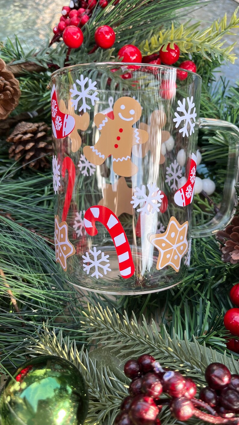 May include: A clear glass mug with a festive Christmas design. The mug features a gingerbread man, candy canes, snowflakes, and stars. The mug is surrounded by a green Christmas tree branch with red berries.