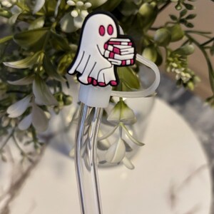 Puede incluir: Una pajita de vidrio transparente con un amuleto de fantasma blanco que sostiene una pila de libros rosas y blancos. El amuleto est&aacute; unido a la pajita con un anillo blanco.